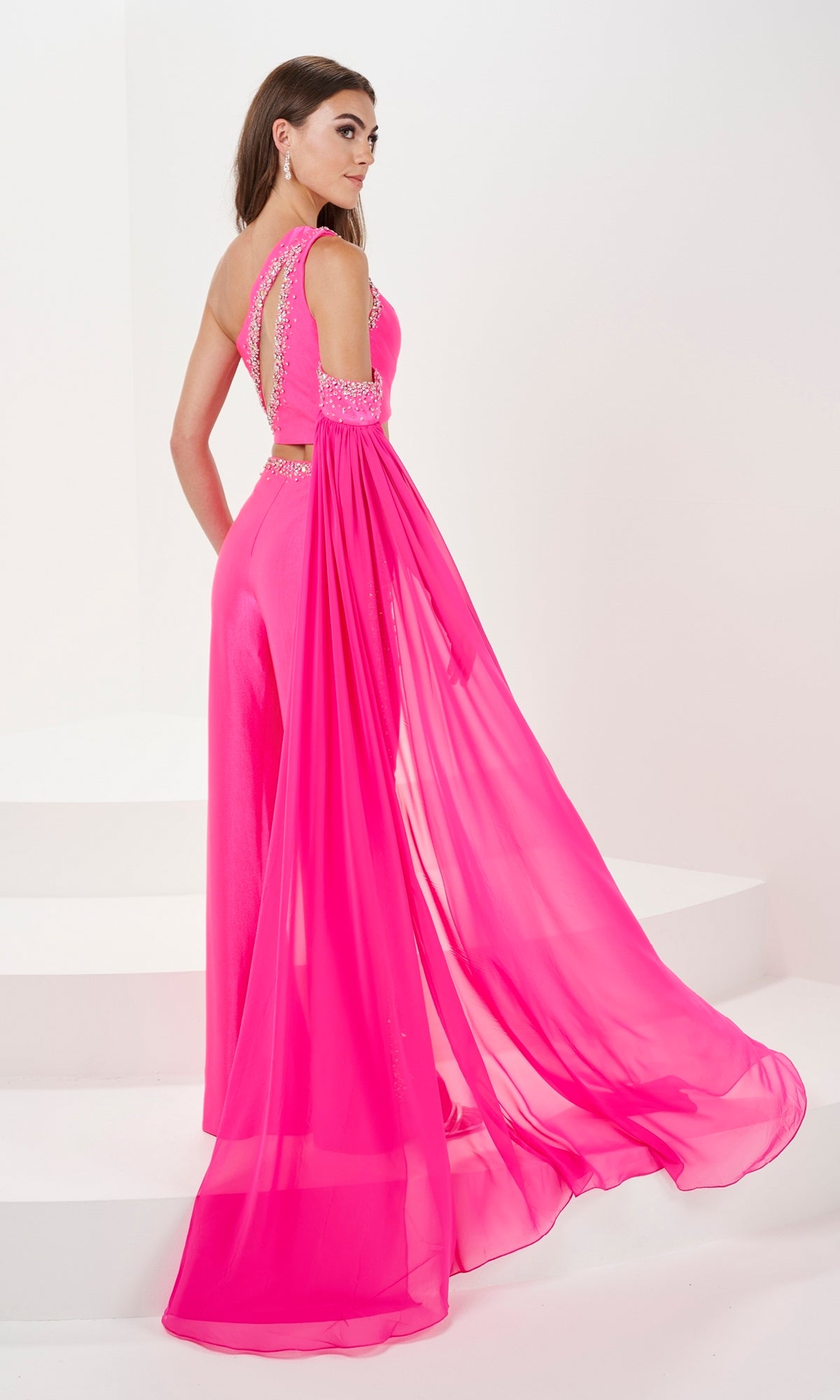 A woman stands elegantly on white steps wearing the Two-Piece One-Shoulder Long Formal Jumpsuit 14165, featuring a bright pink hue, beaded details, and a flowing sheer cape.